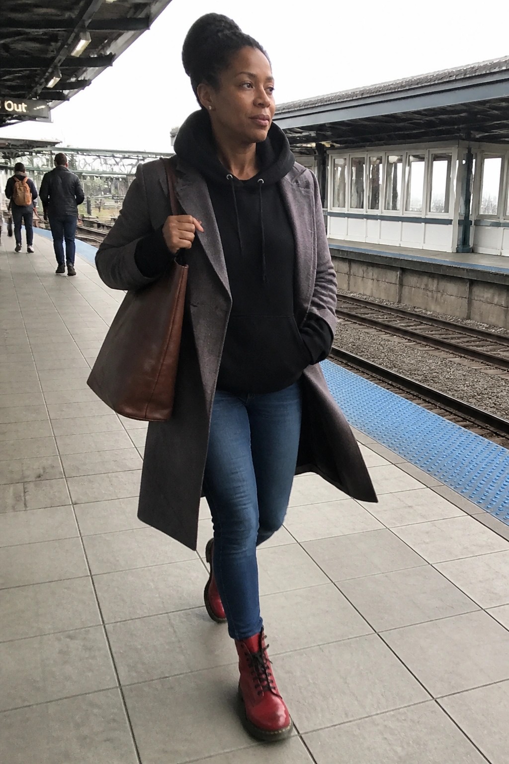 Woman in gray coat over black hoodie, blue jeans and red boots holds brown bag on platform