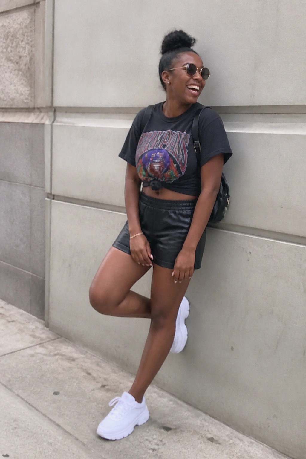 Woman with high bun smiling against stone wall in black t-shirt and leather shorts.
