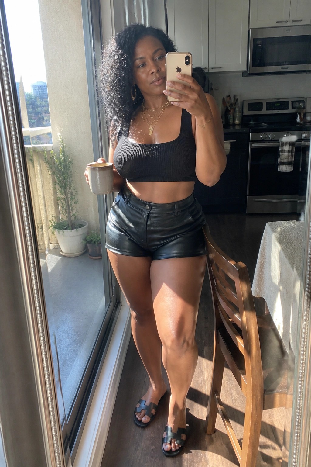 Woman in black crop top and leather shorts holds phone and mug in kitchen