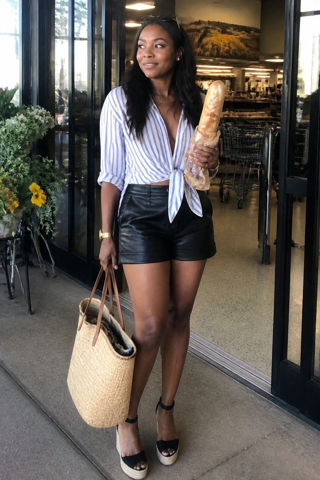 Woman in striped tie-front shirt and leather shorts holding baguette and straw tote bag