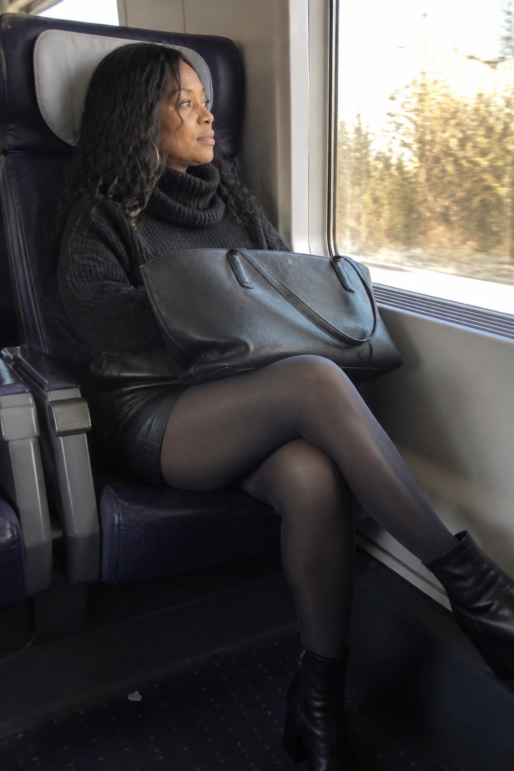 Woman wearing black leather shorts, black chunky turtleneck sweater, sheer black tights, and black ankle boots while holding a large black leather tote bag.