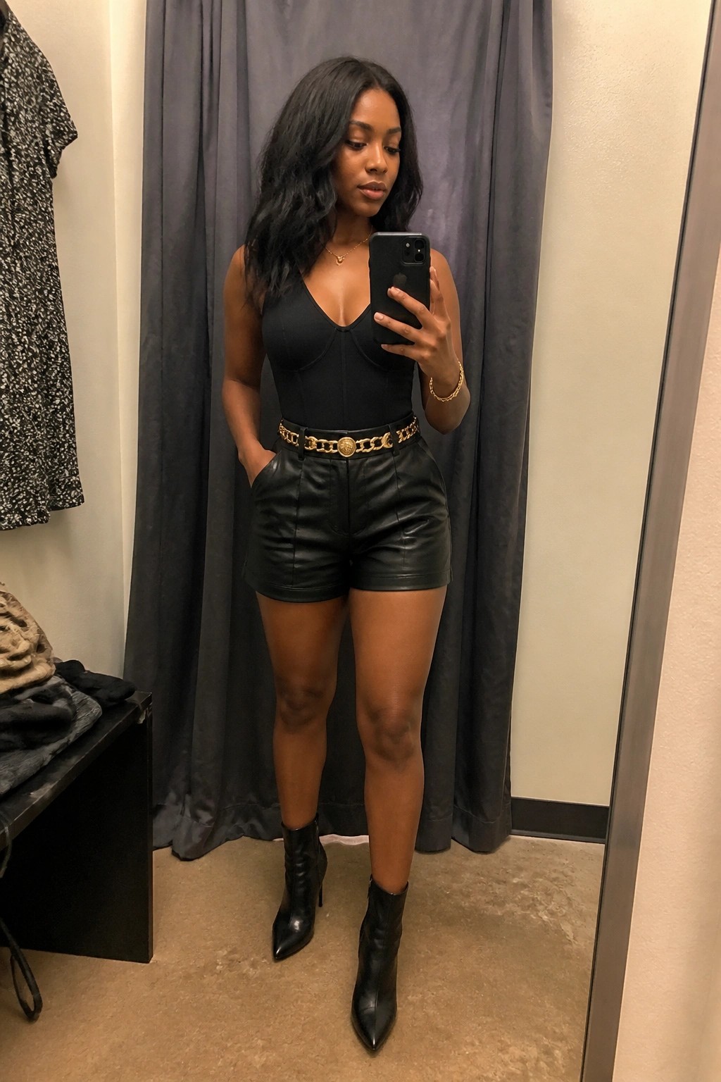 Woman wearing black leather shorts, black sleeveless corset bodysuit, gold chain belt, black pointed ankle boots, and gold jewelry while taking a mirror selfie in a fitting room.
