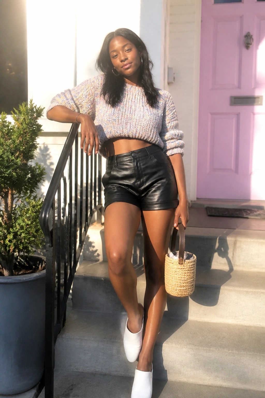 Woman in colorful knit crop top and black leather shorts holding woven bag on steps