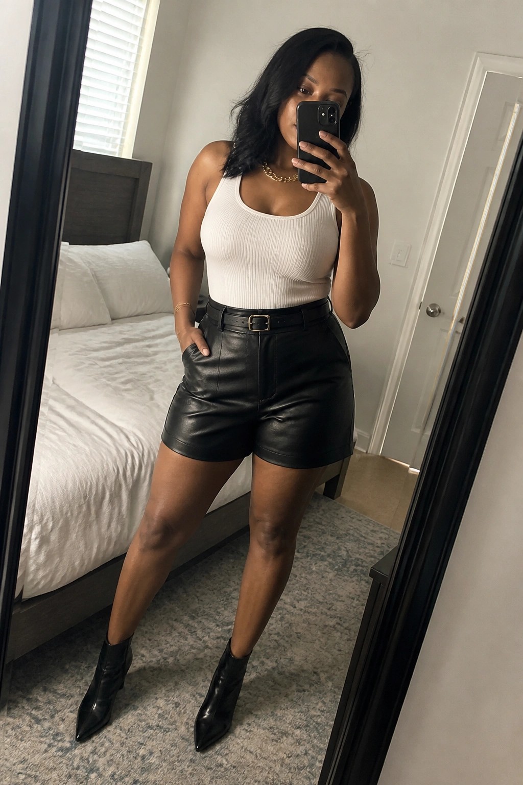 Woman in white tank top and black leather shorts taking bedroom mirror selfie