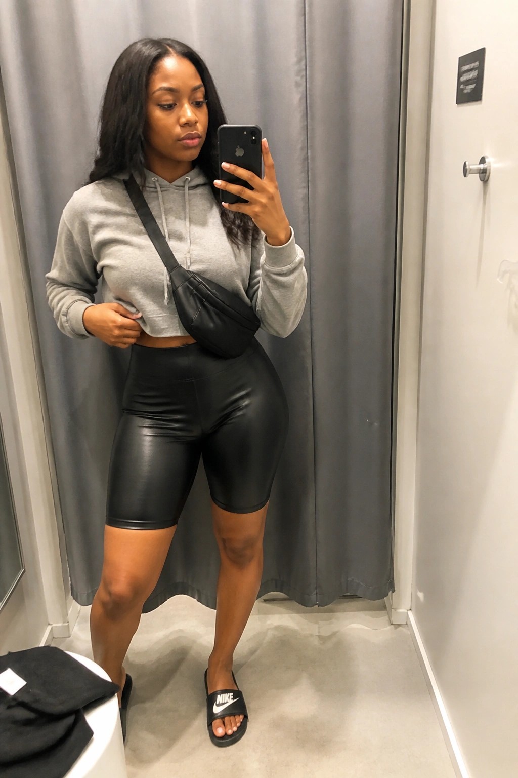 Woman in a fitting room mirror selfie wearing a cropped gray hoodie, black leather biker shorts, black crossbody bag, and black Nike slides.