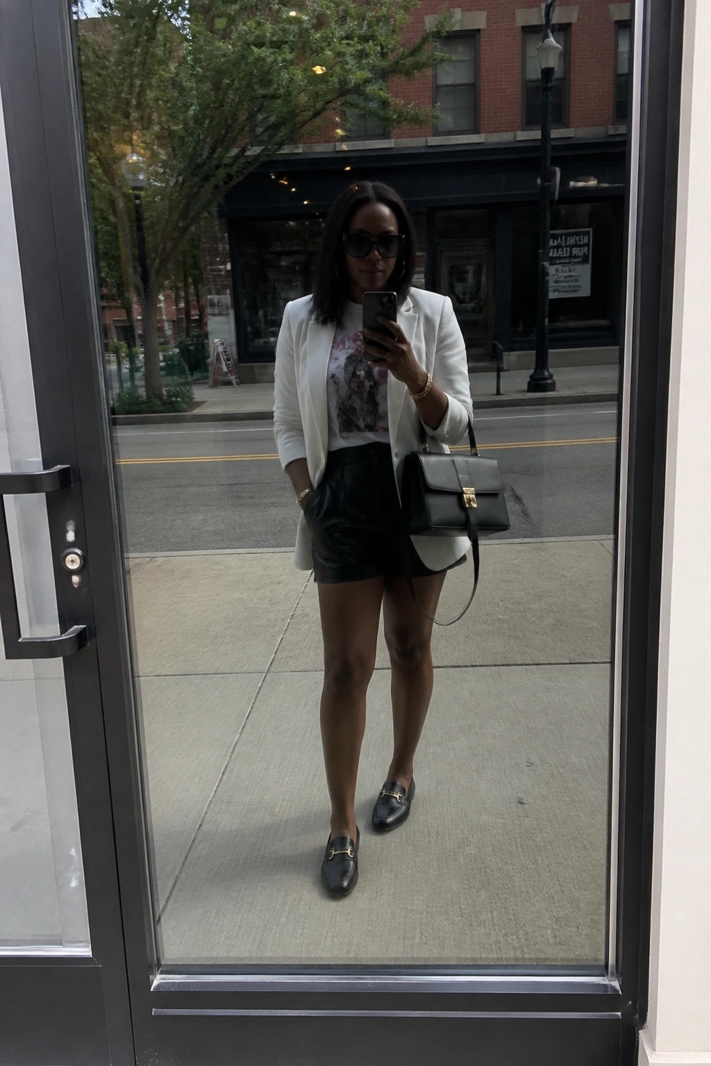 Woman in white blazer and black leather shorts taking selfie in glass reflection