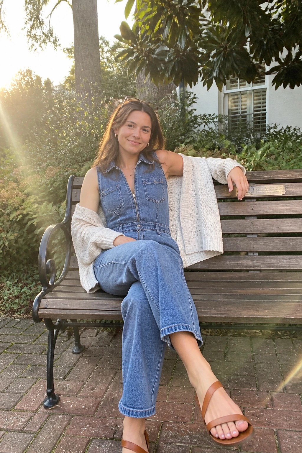 Young woman in blue denim jumpsuit and white cardigan relaxes on wooden park bench.