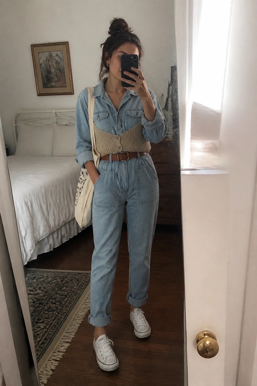 Woman wearing light wash denim jumpsuit with beige knit vest, brown belt, white sneakers, and tote bag in mirror selfie.
