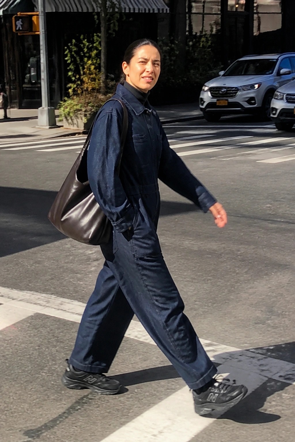 Woman in navy denim jumpsuit crosses urban street with brown leather bag.