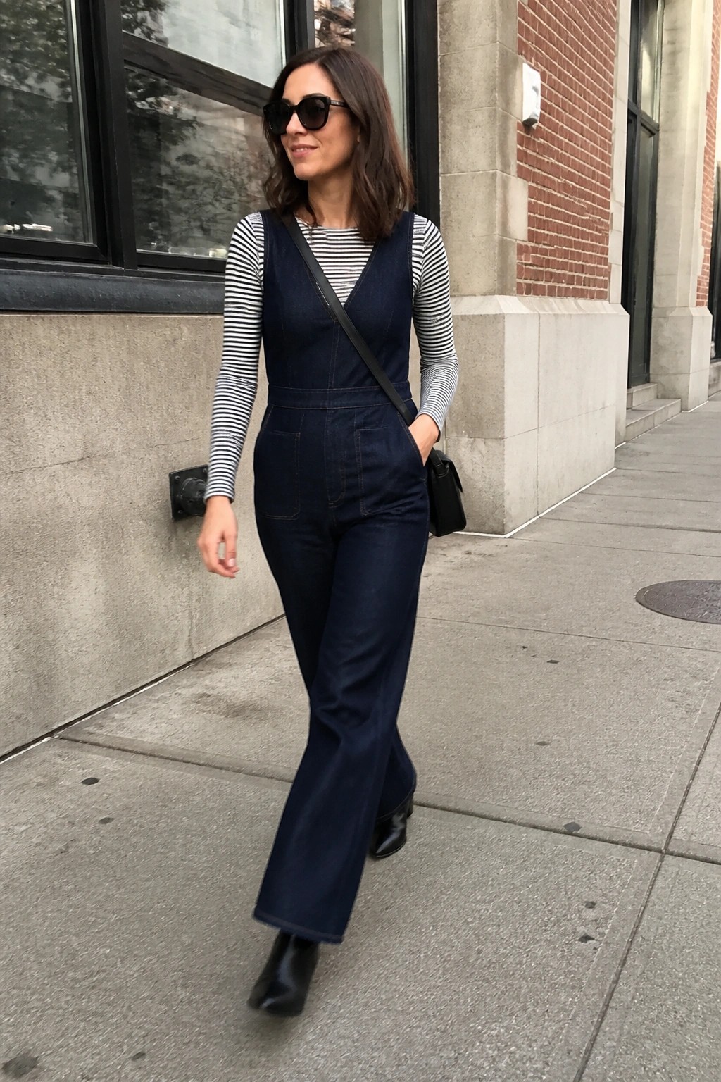 Woman in dark blue denim jumpsuit over black and white striped shirt with black ankle boots and crossbody bag walking on sidewalk.