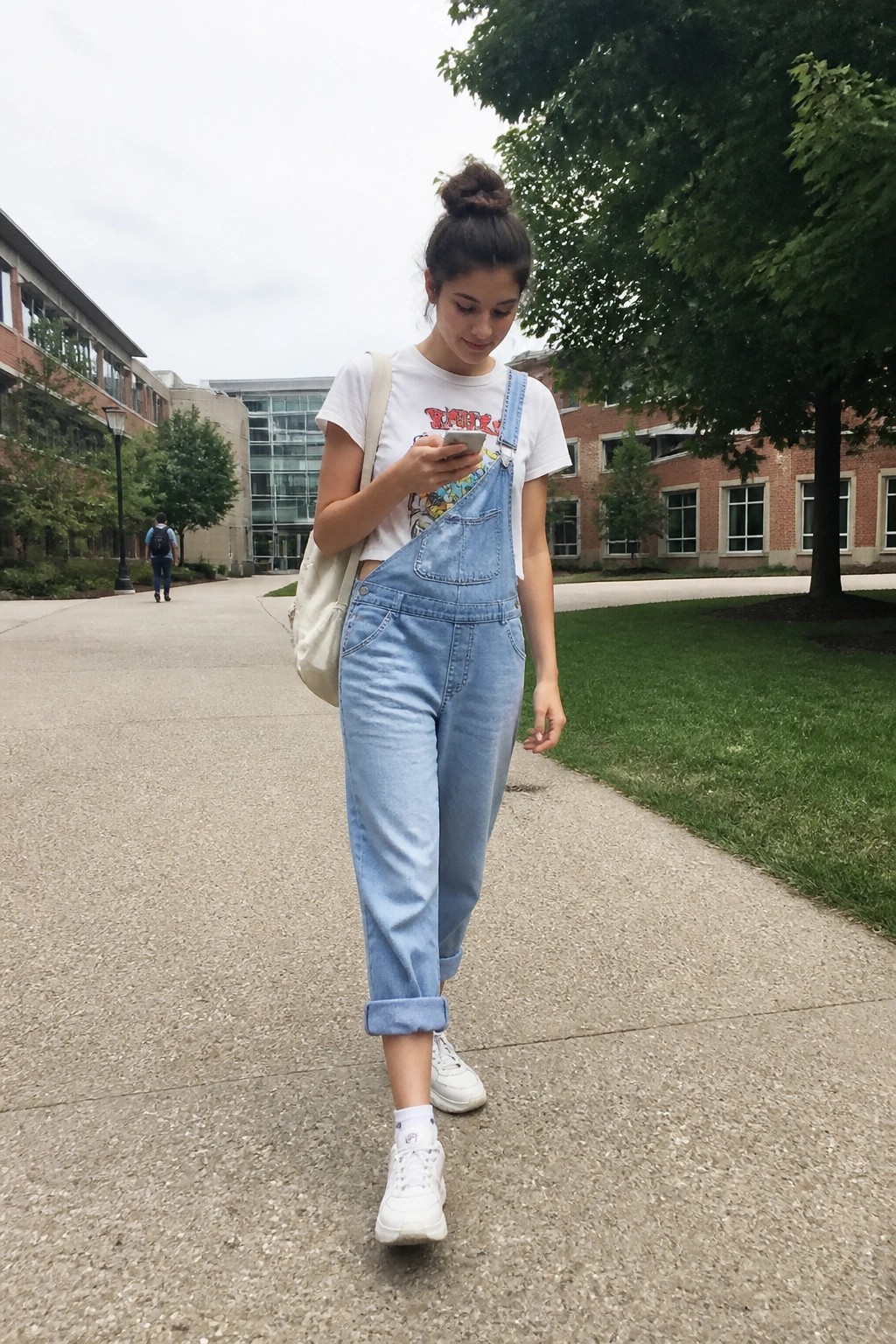 Young woman in light blue overalls checks phone on campus walkway