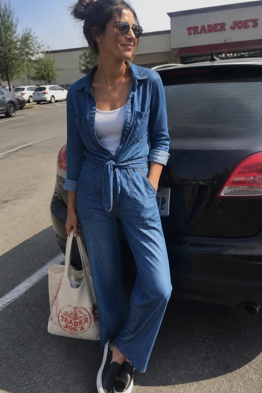 Woman in blue denim jumpsuit holding Trader Joe's tote bag by car