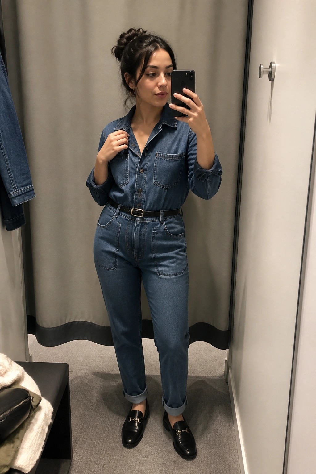 Woman in blue denim jumpsuit taking mirror selfie in fitting room with black loafers