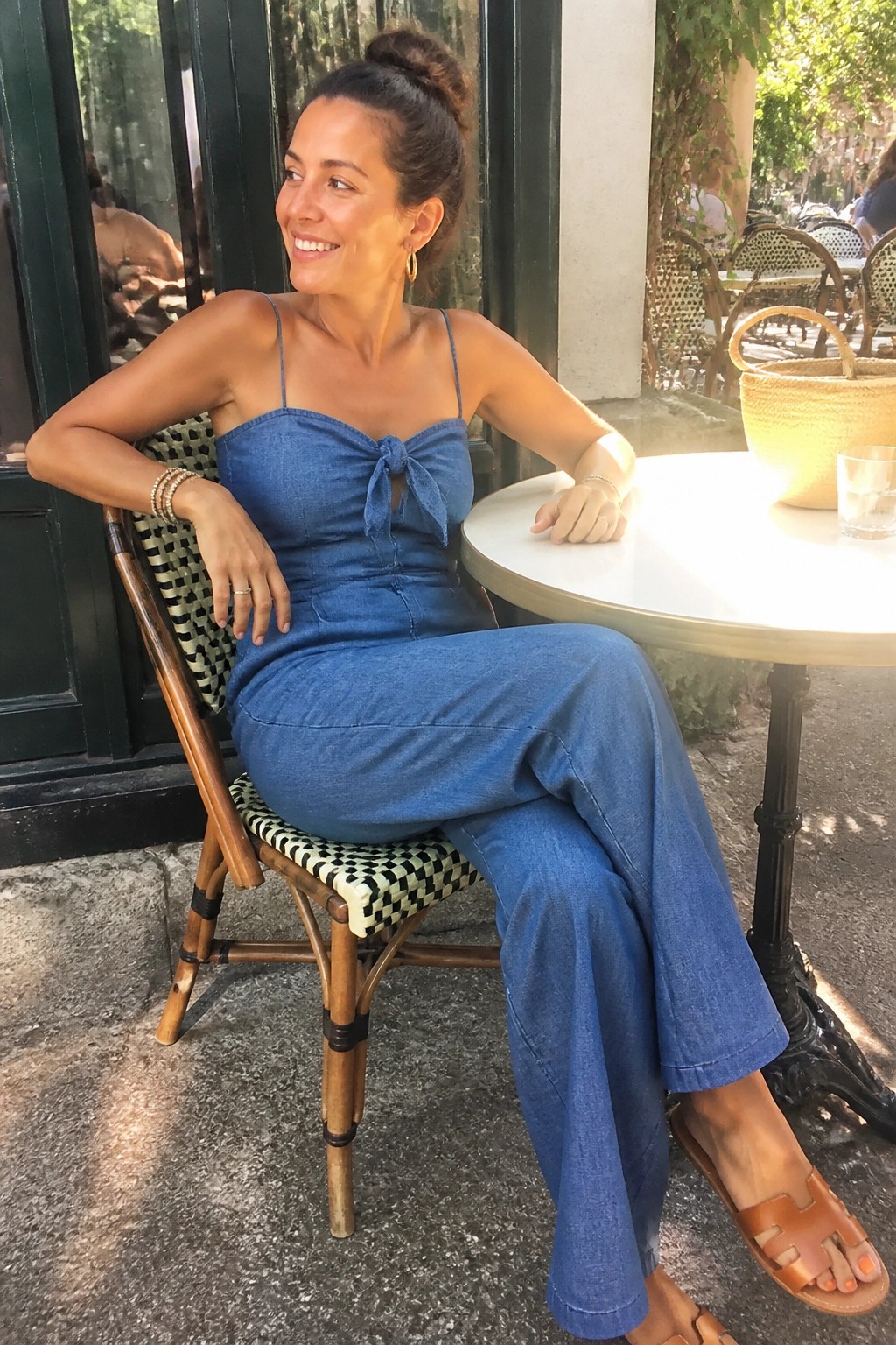 Woman in blue denim jumpsuit seated at sunny outdoor cafe table