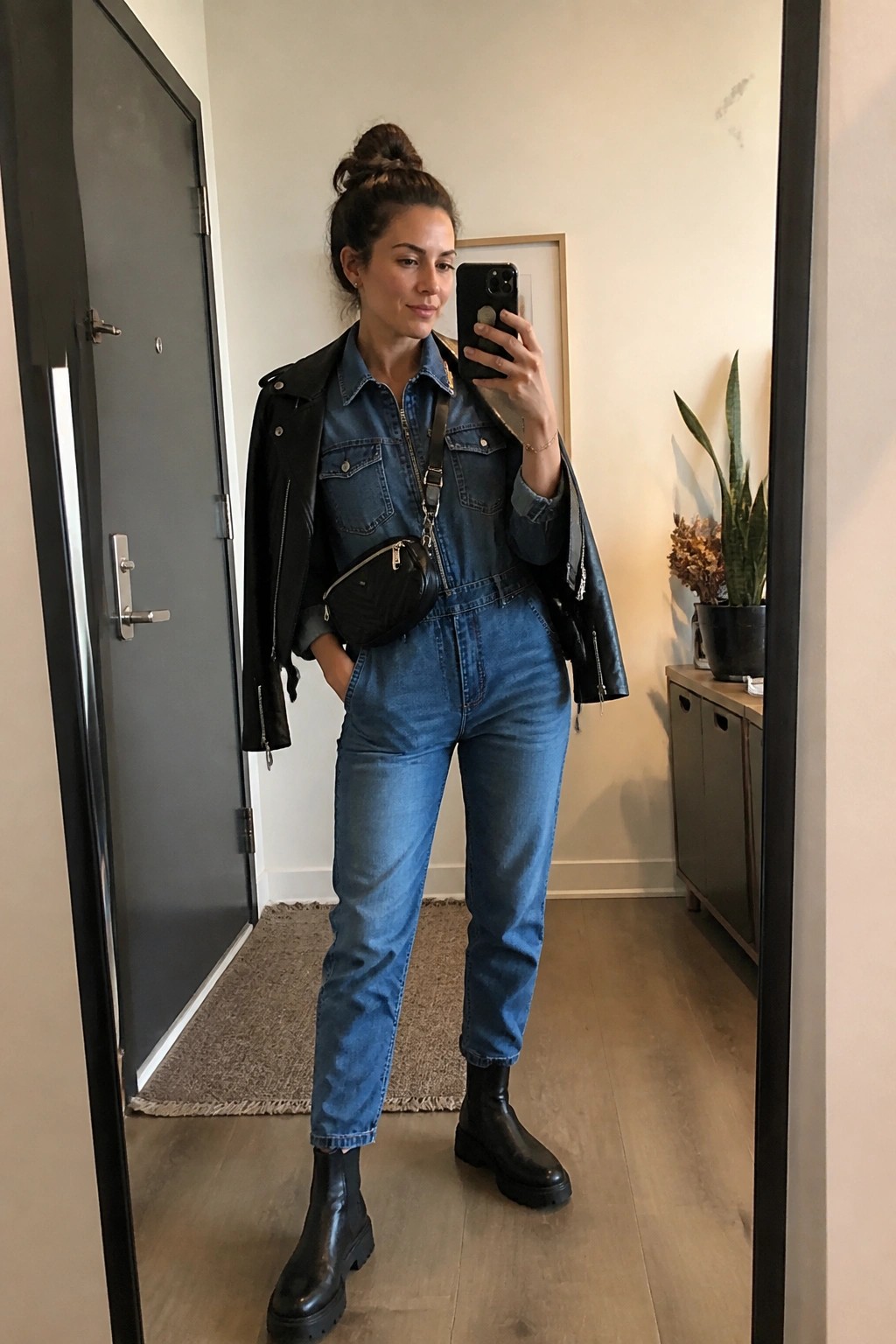 Woman wearing a blue denim jumpsuit with a black leather jacket draped over her shoulders, black chunky boots, and a small black crossbody bag while taking a mirror selfie.