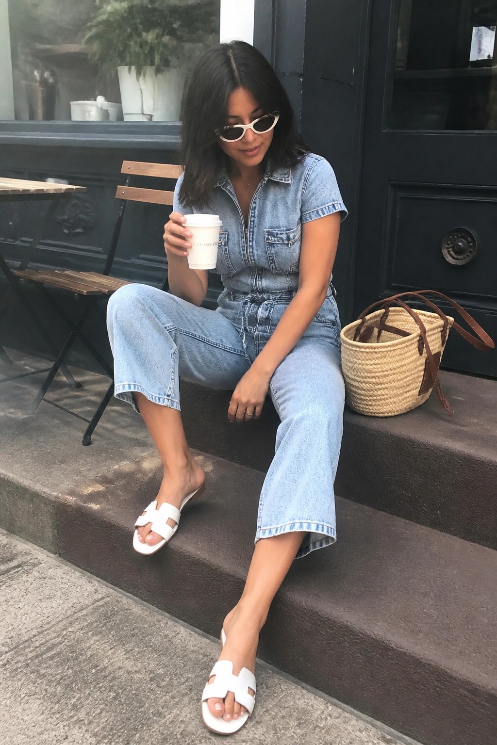 Woman wearing light blue short sleeve denim jumpsuit with wide cropped legs, white slide sandals featuring an H cutout, white coffee cup in hand, and woven basket bag nearby on steps.