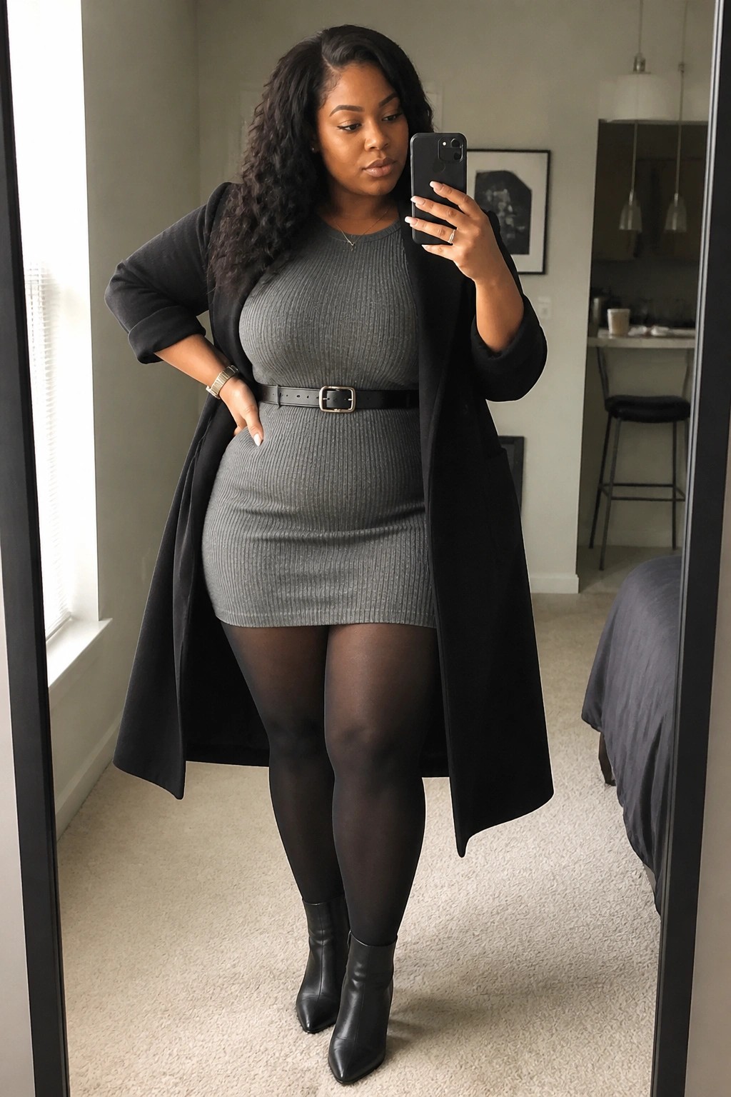 A plus size Black woman in a gray ribbed mini dress cinched with a black belt, layered under an open black coat, paired with black tights and pointed ankle boots while holding a phone for a mirror selfie.