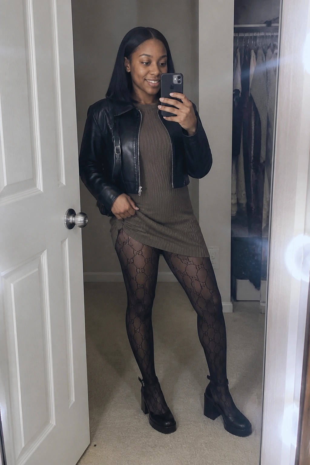 Woman in taupe ribbed dress and black leather jacket taking mirror selfie with phone.