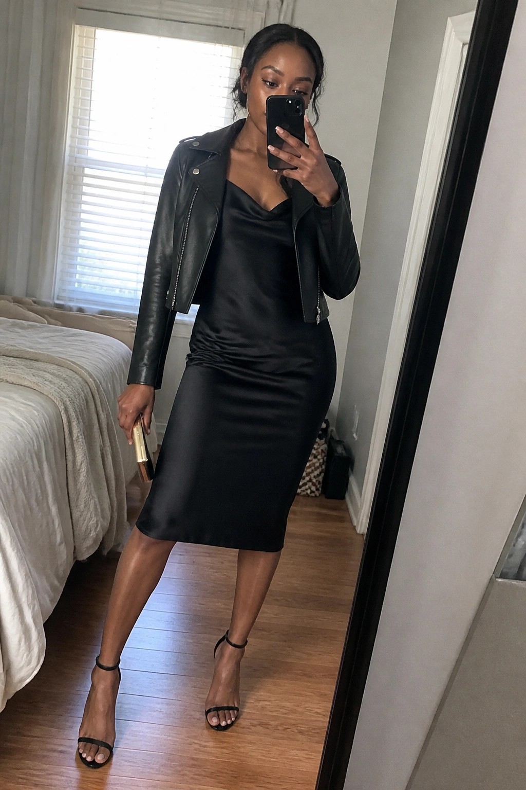 Woman in black leather jacket and satin dress taking mirror selfie in bedroom