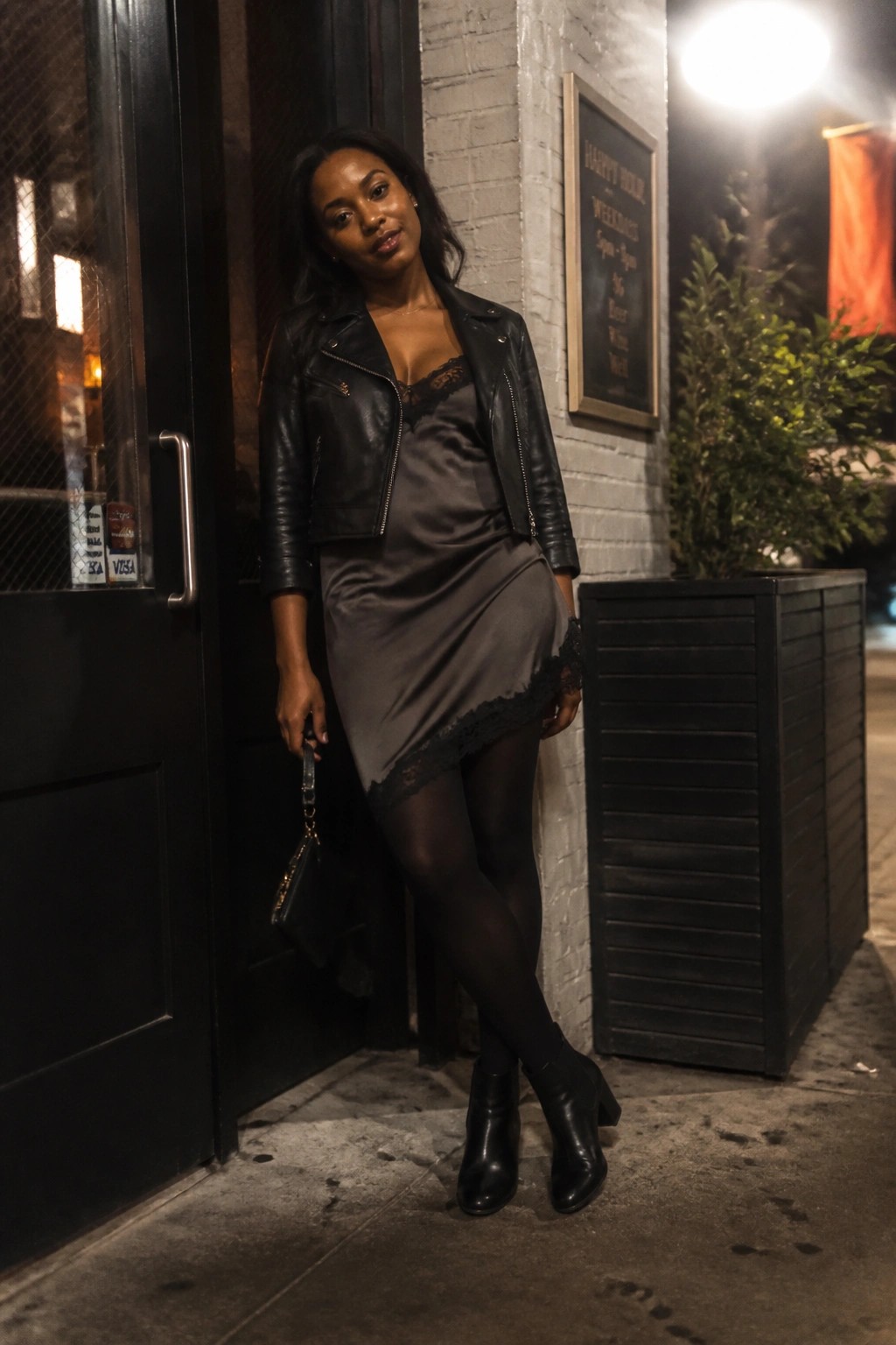 Woman in black leather jacket and gray lace dress posing outside at night.
