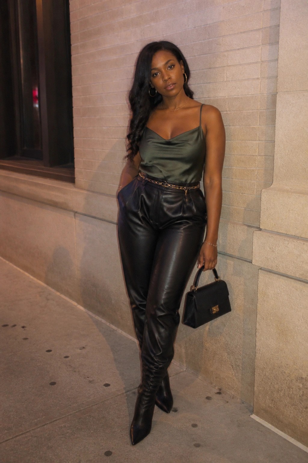 Woman in green satin camisole and black leather pants holding handbag by brick wall
