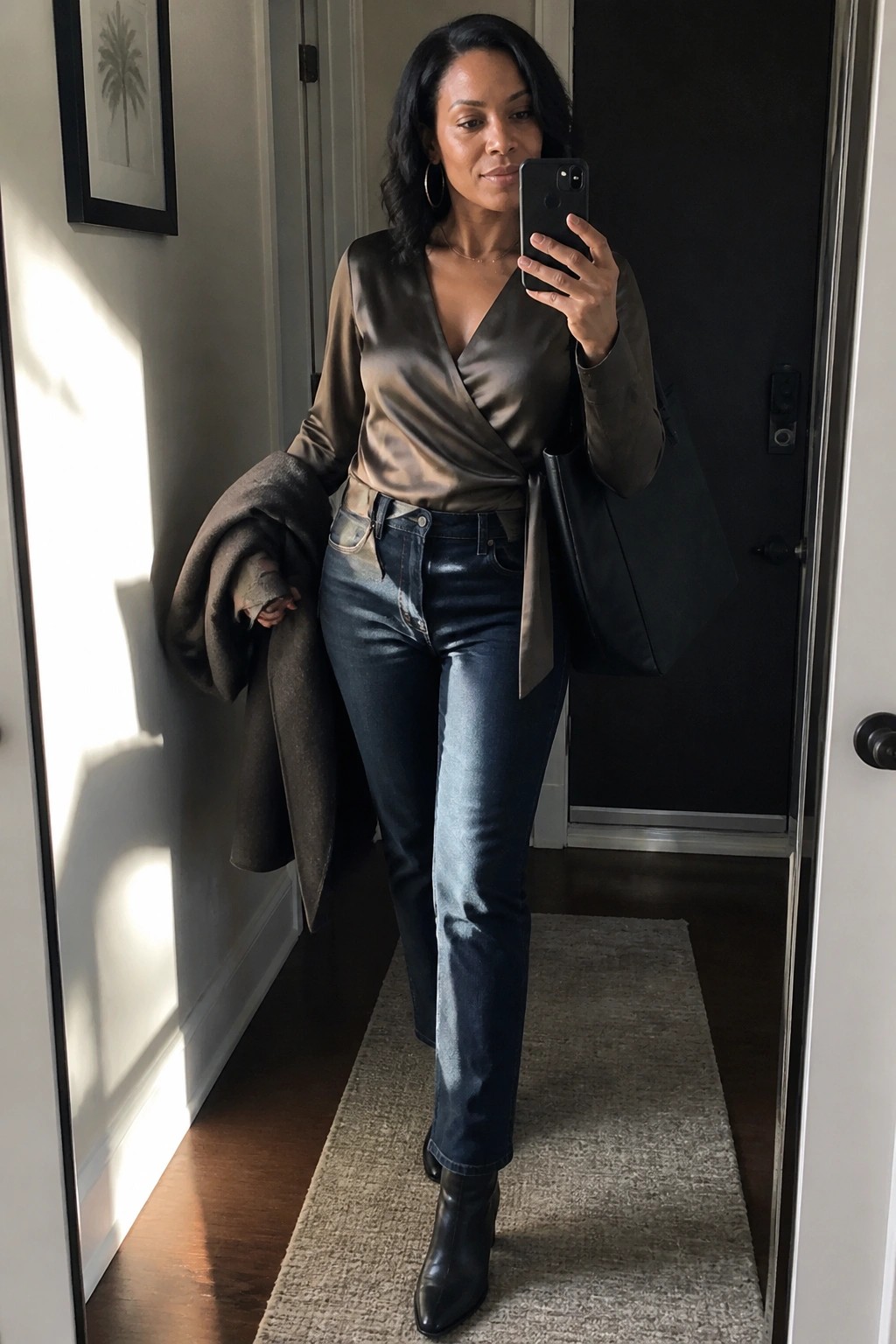 Woman in brown satin wrap top and jeans taking mirror selfie in hallway