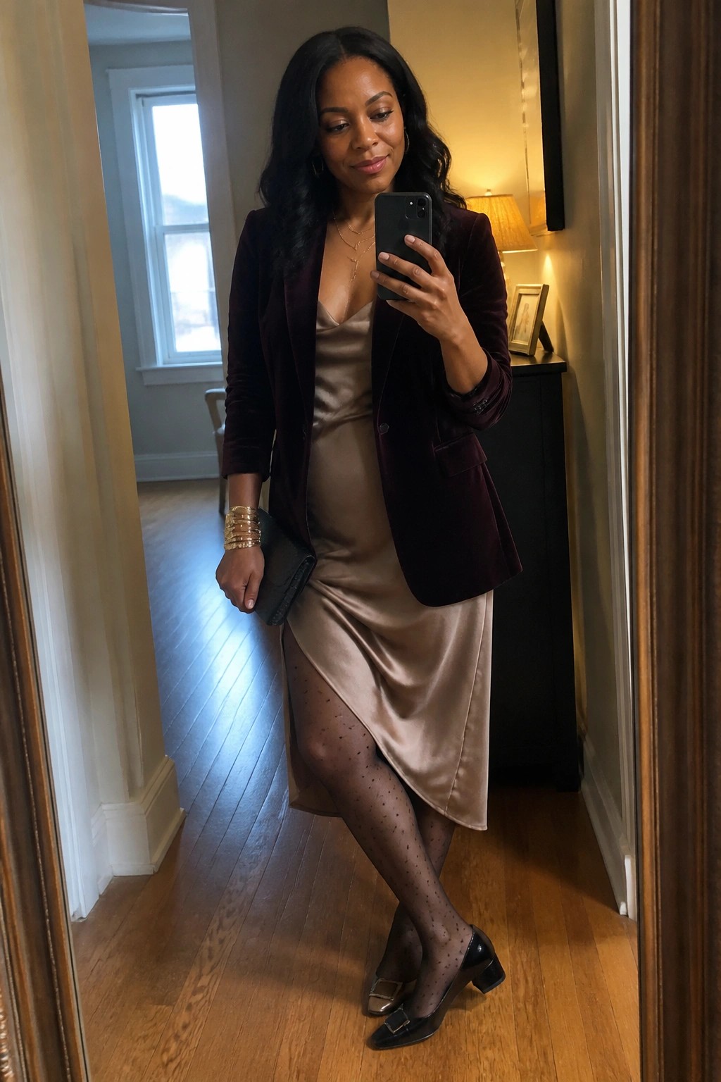 A woman wearing a burgundy velvet blazer over a champagne satin slip dress with polka dot tights and black heels, holding a phone for a mirror selfie.