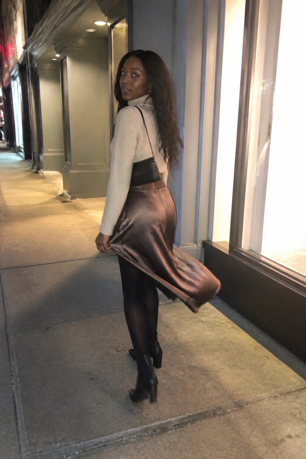 Woman in white turtleneck and flowing brown satin skirt on nighttime sidewalk