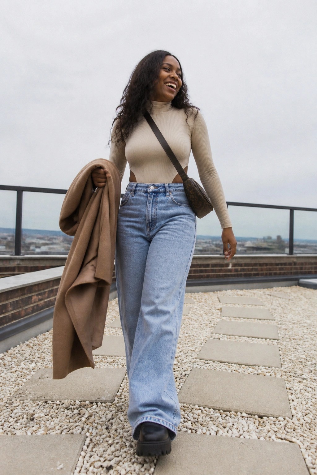 Smiling woman in beige cutout top and blue jeans holding brown coat on rooftop.