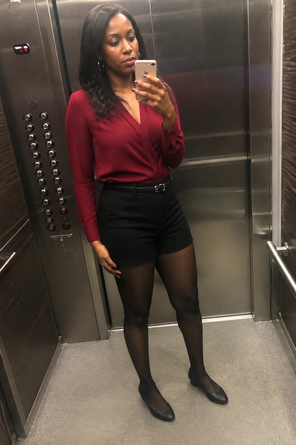 Woman in red blouse and black shorts taking selfie in elevator