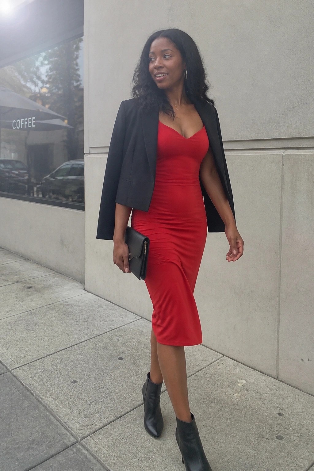 Woman in red midi dress with black blazer over shoulders holding clutch on sidewalk