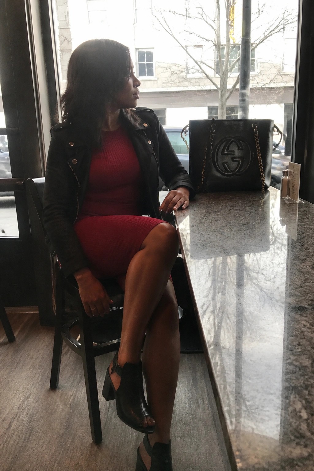 Woman wearing a red ribbed knit dress under a black leather jacket paired with black open-toe block heel booties and a large black Gucci tote bag placed on the counter beside her.