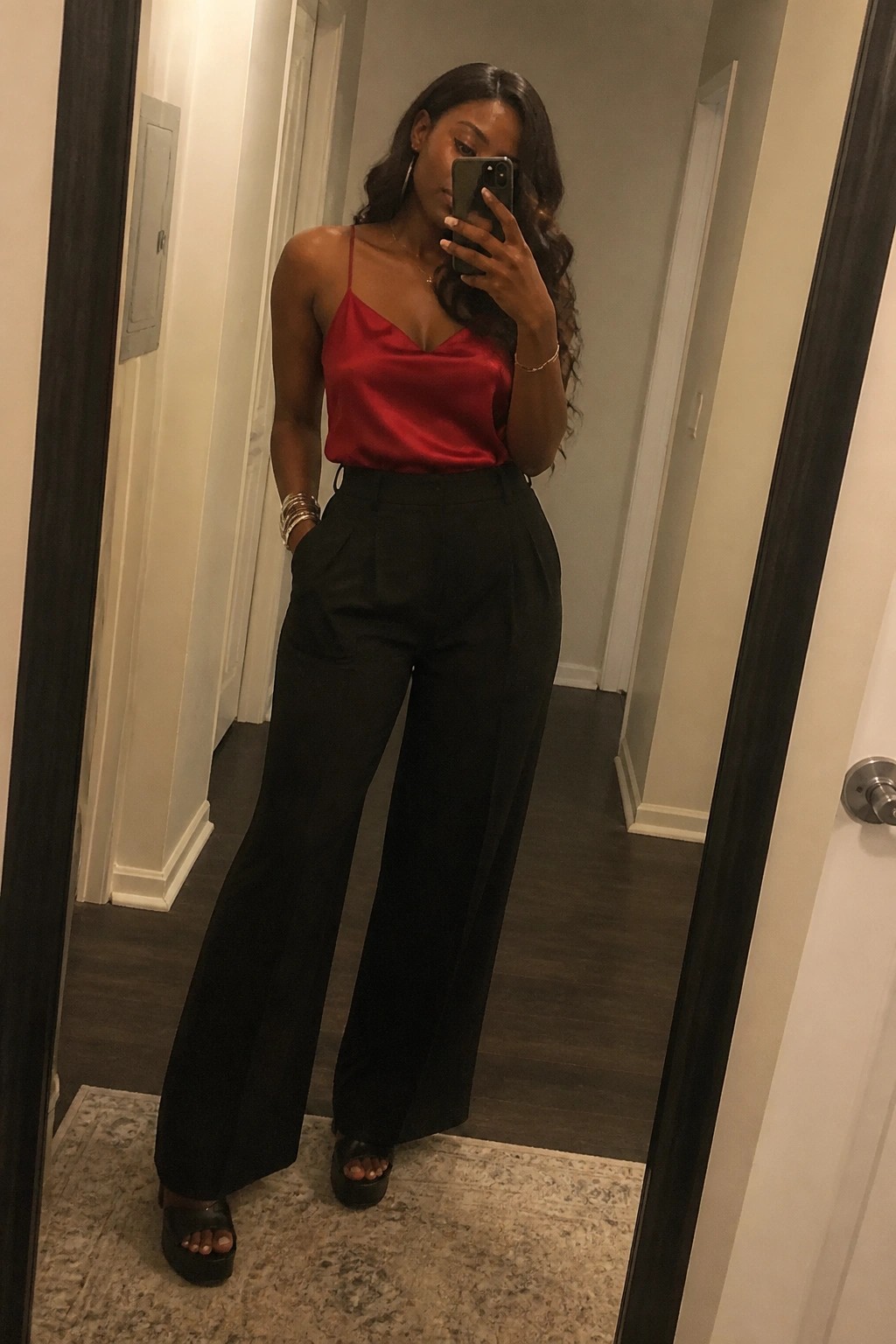 Woman wearing a red satin camisole top with black wide-leg pleated trousers and black platform sandals while taking a mirror selfie in a hallway.