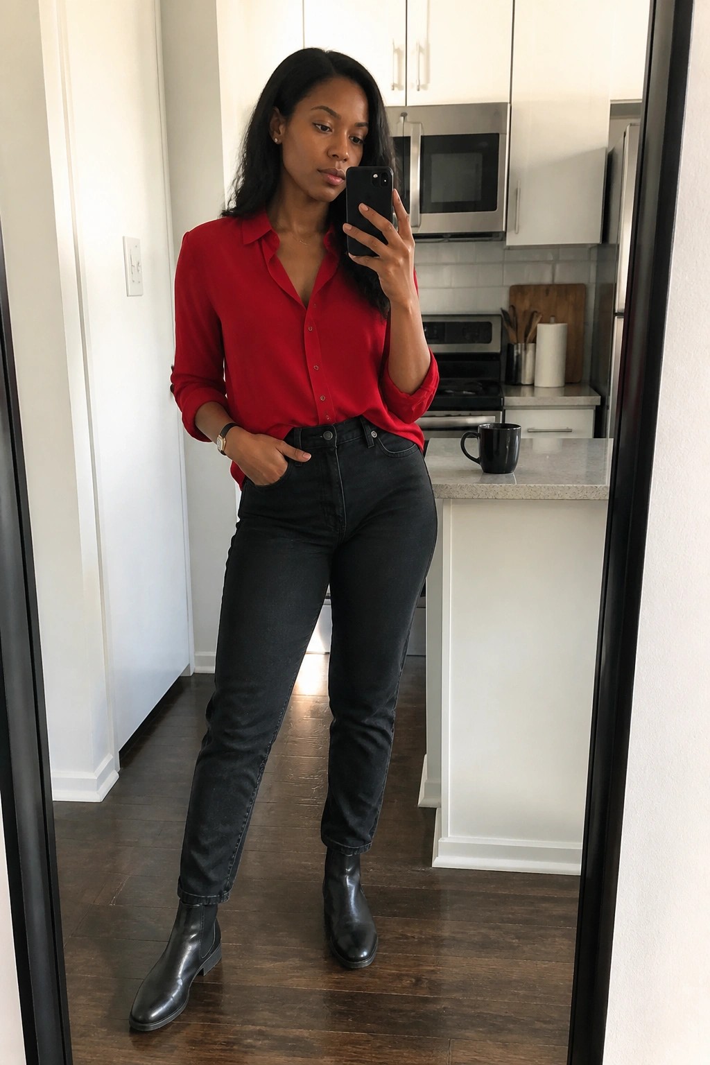 Woman in red shirt and black jeans taking mirror selfie in kitchen