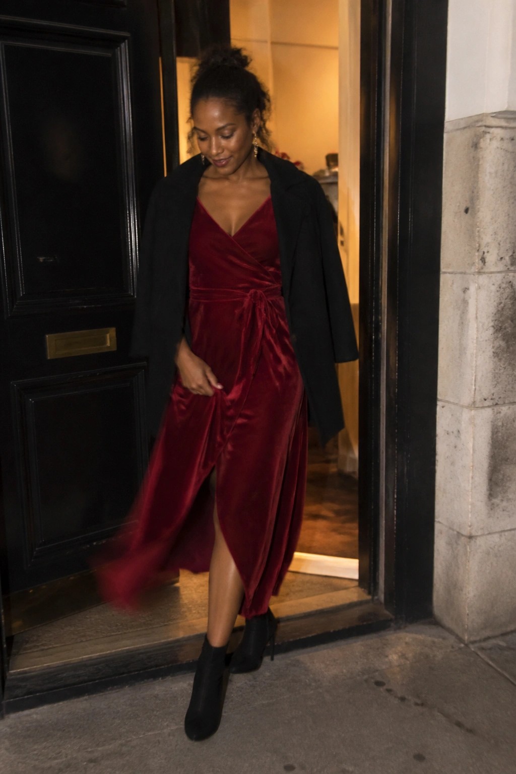 Woman in red velvet dress and black coat standing in doorway at night