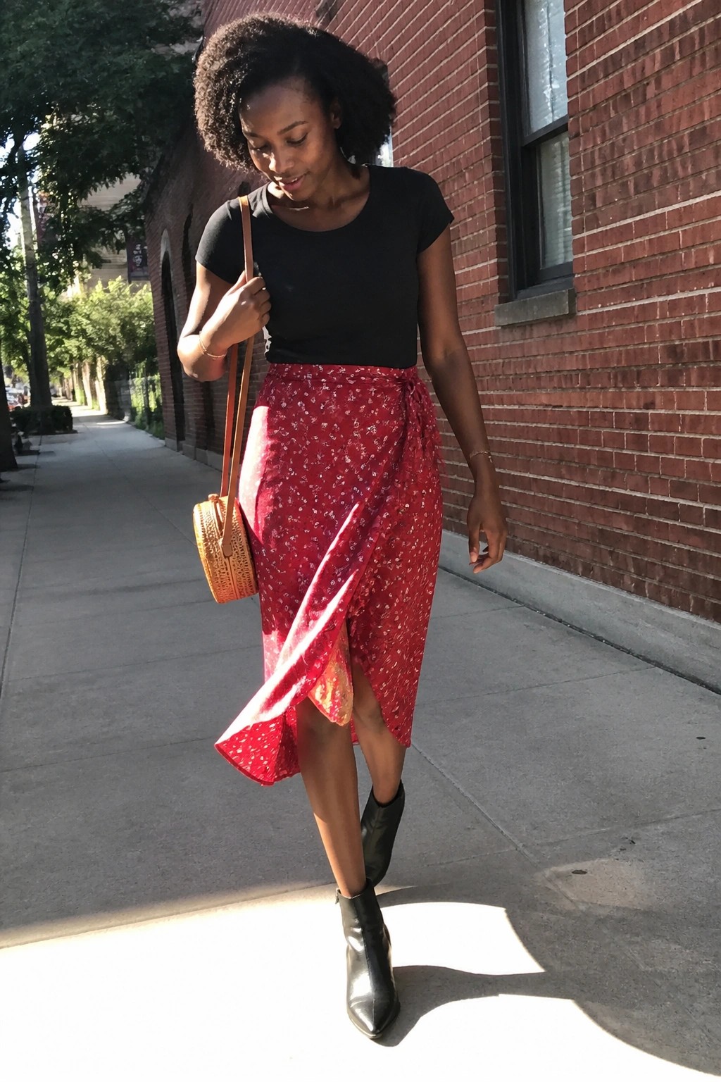 Woman in black top and red floral wrap skirt carrying round bag on sidewalk.