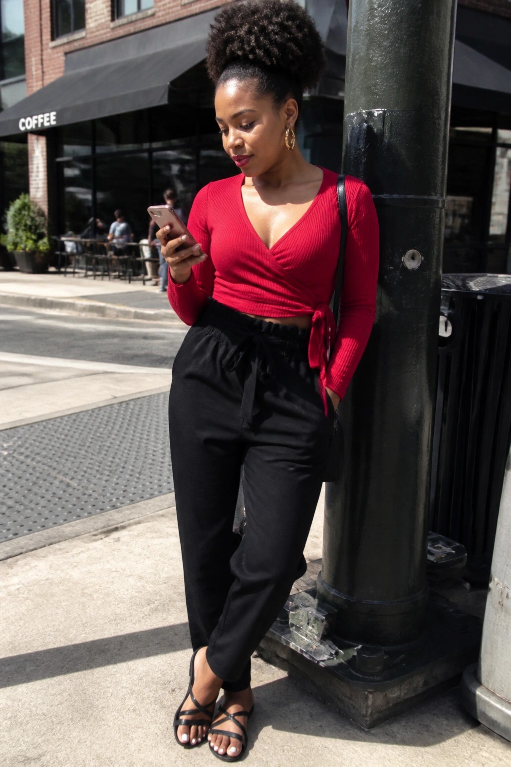 Black woman in red wrap crop top and black drawstring pants with strappy sandals holding a phone outdoors