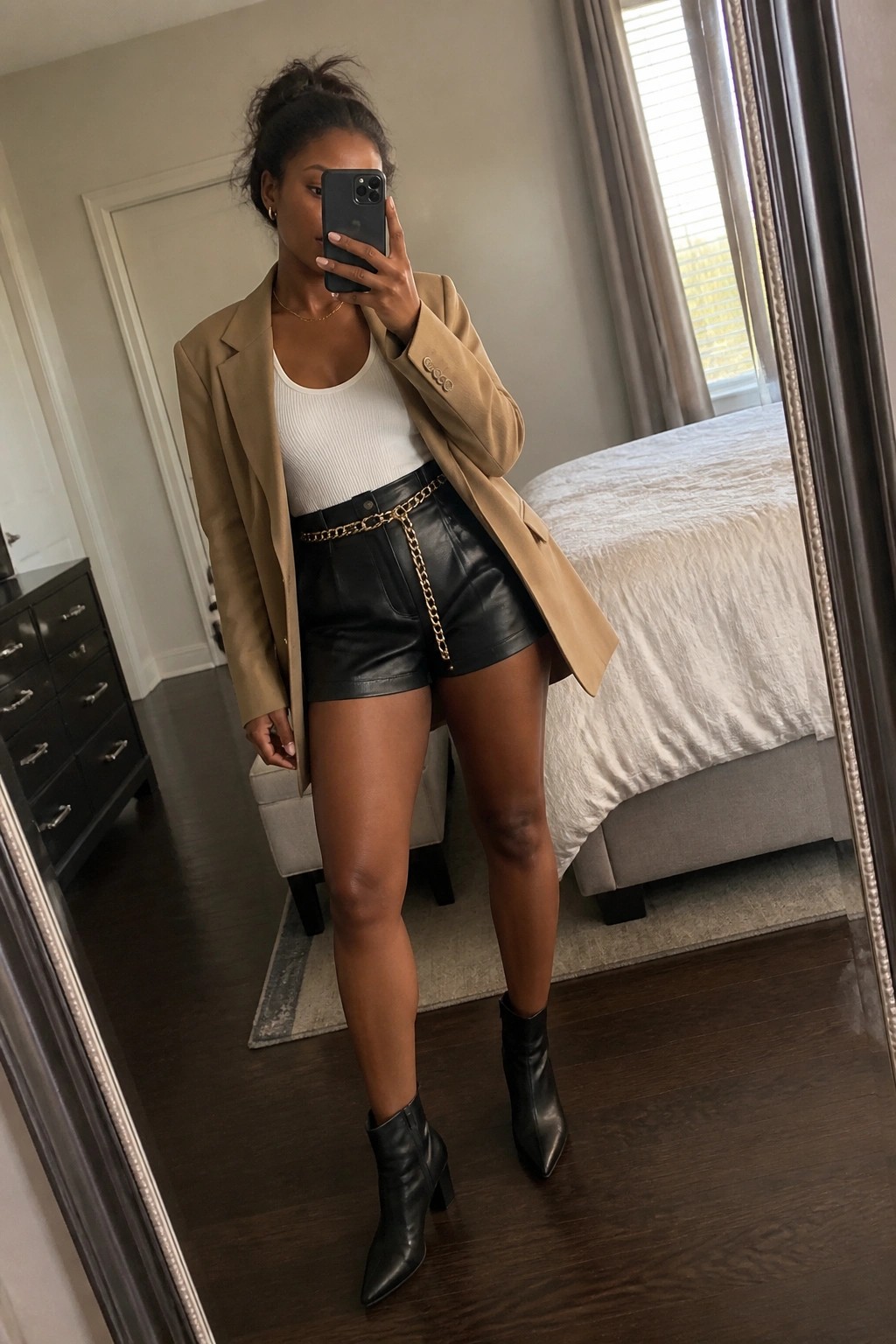 Woman in tan blazer, white tank, black leather shorts with gold chain, taking bedroom mirror selfie