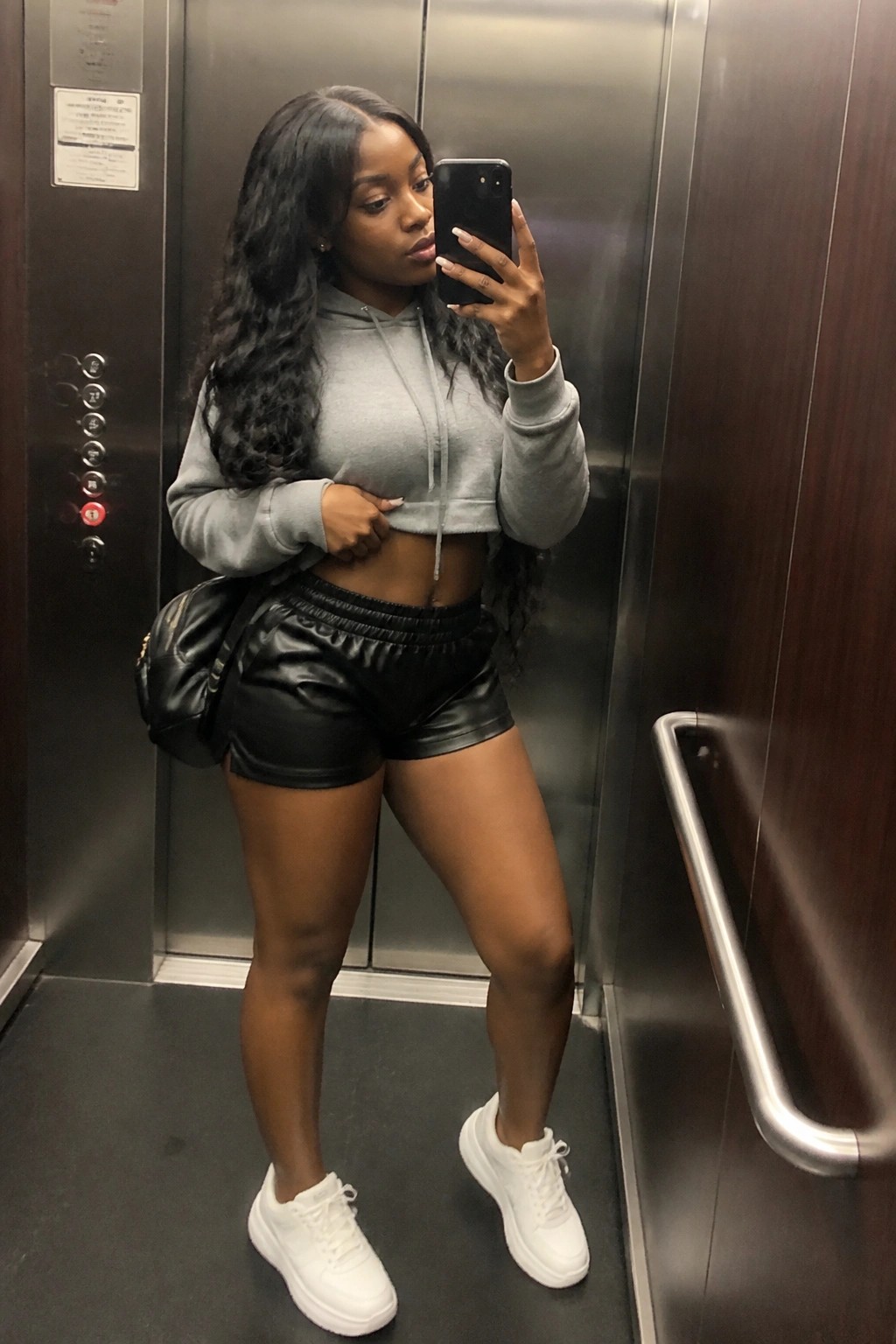 Woman in gray cropped hoodie and black shorts taking selfie in elevator