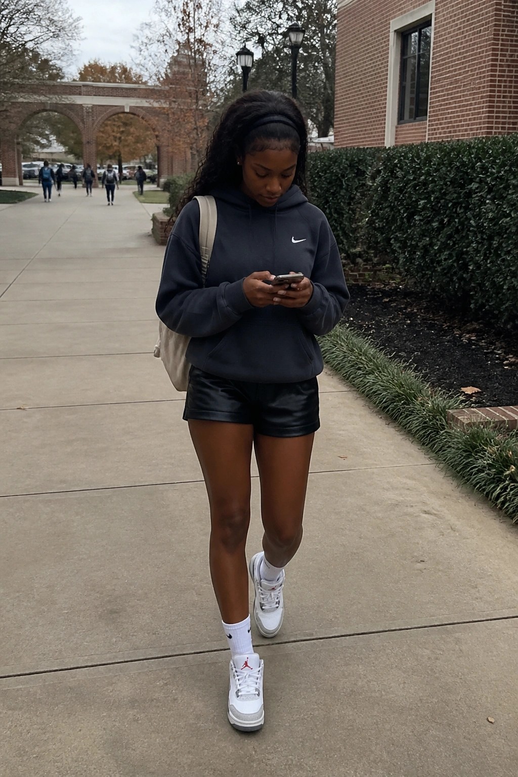 Woman in gray Nike hoodie and black shorts checks phone on campus path