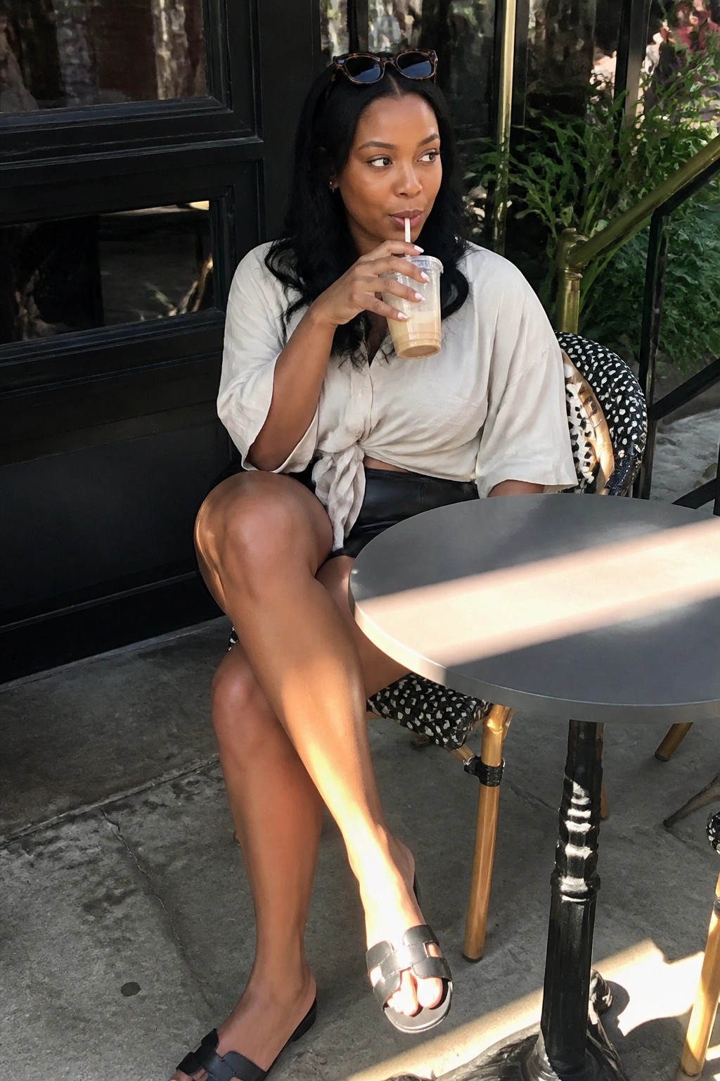 Woman in beige blouse and black skirt sips iced drink at sunny outdoor table