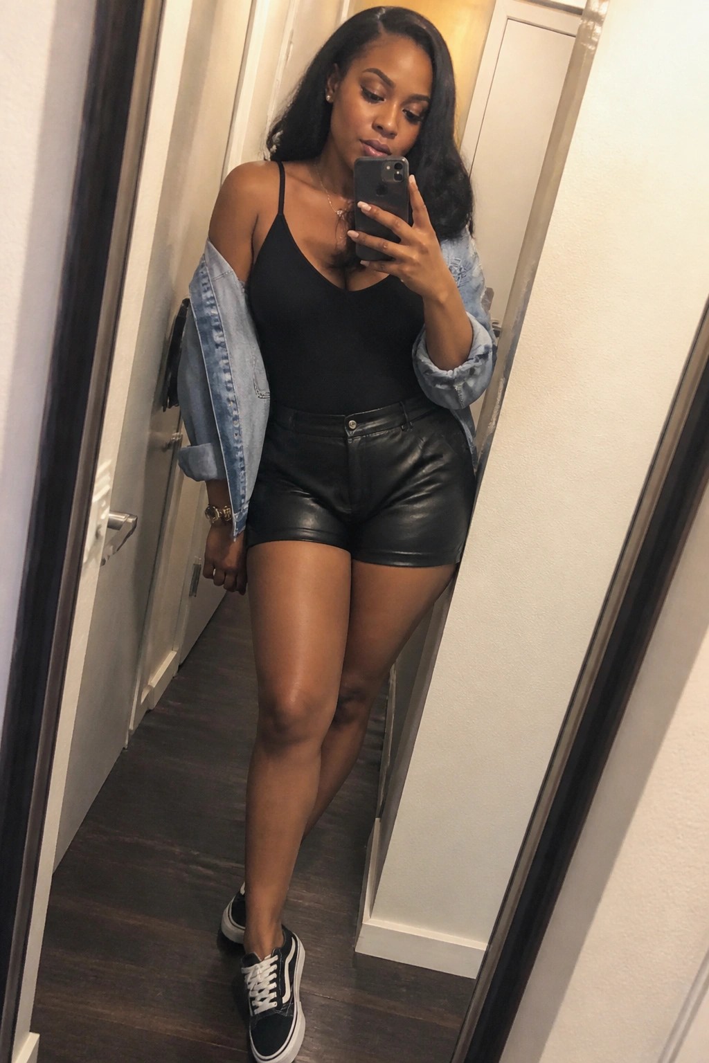Woman in black bodysuit and leather shorts with denim jacket taking mirror selfie