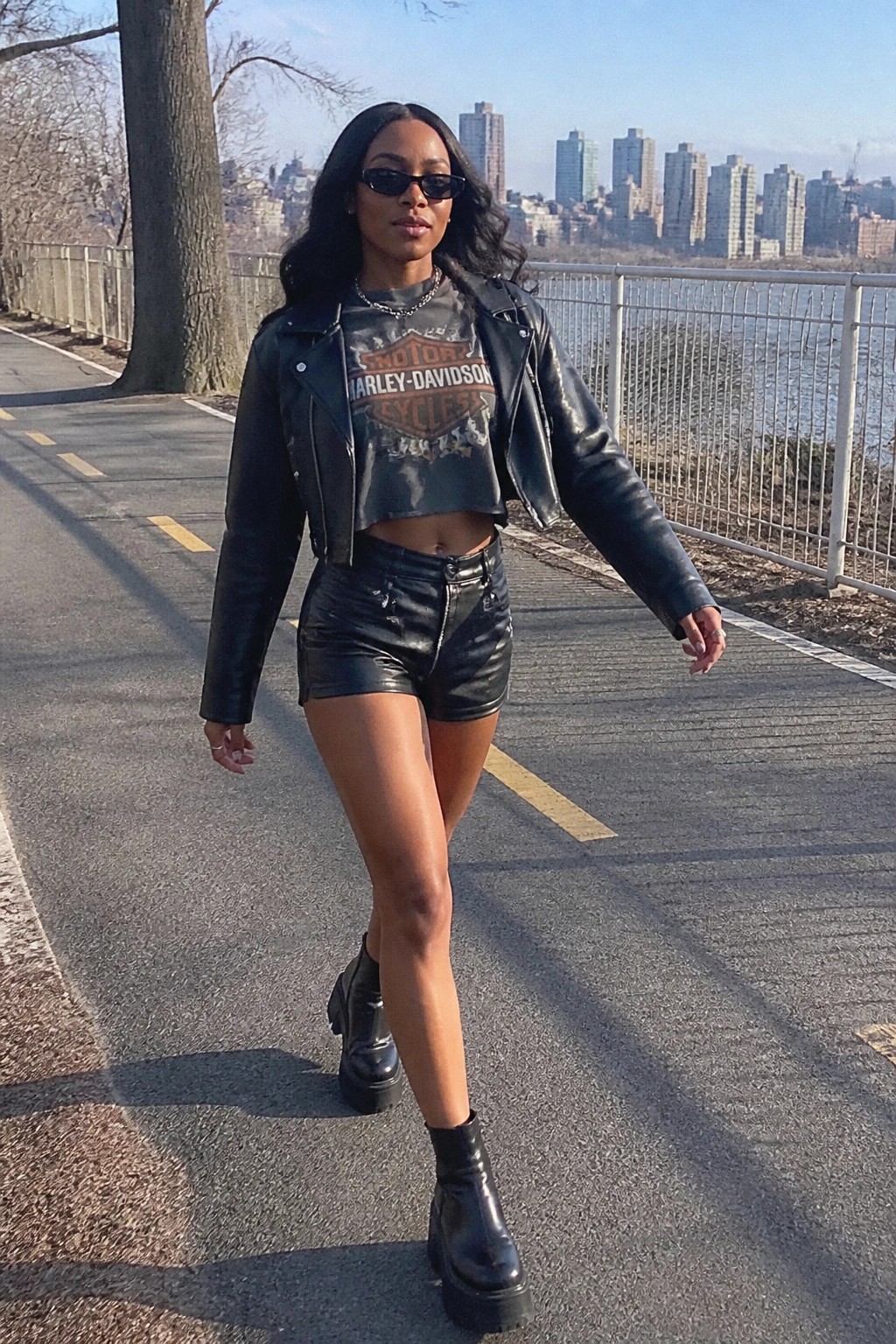 Woman in black leather jacket, Harley-Davidson crop top, and shorts on city path.