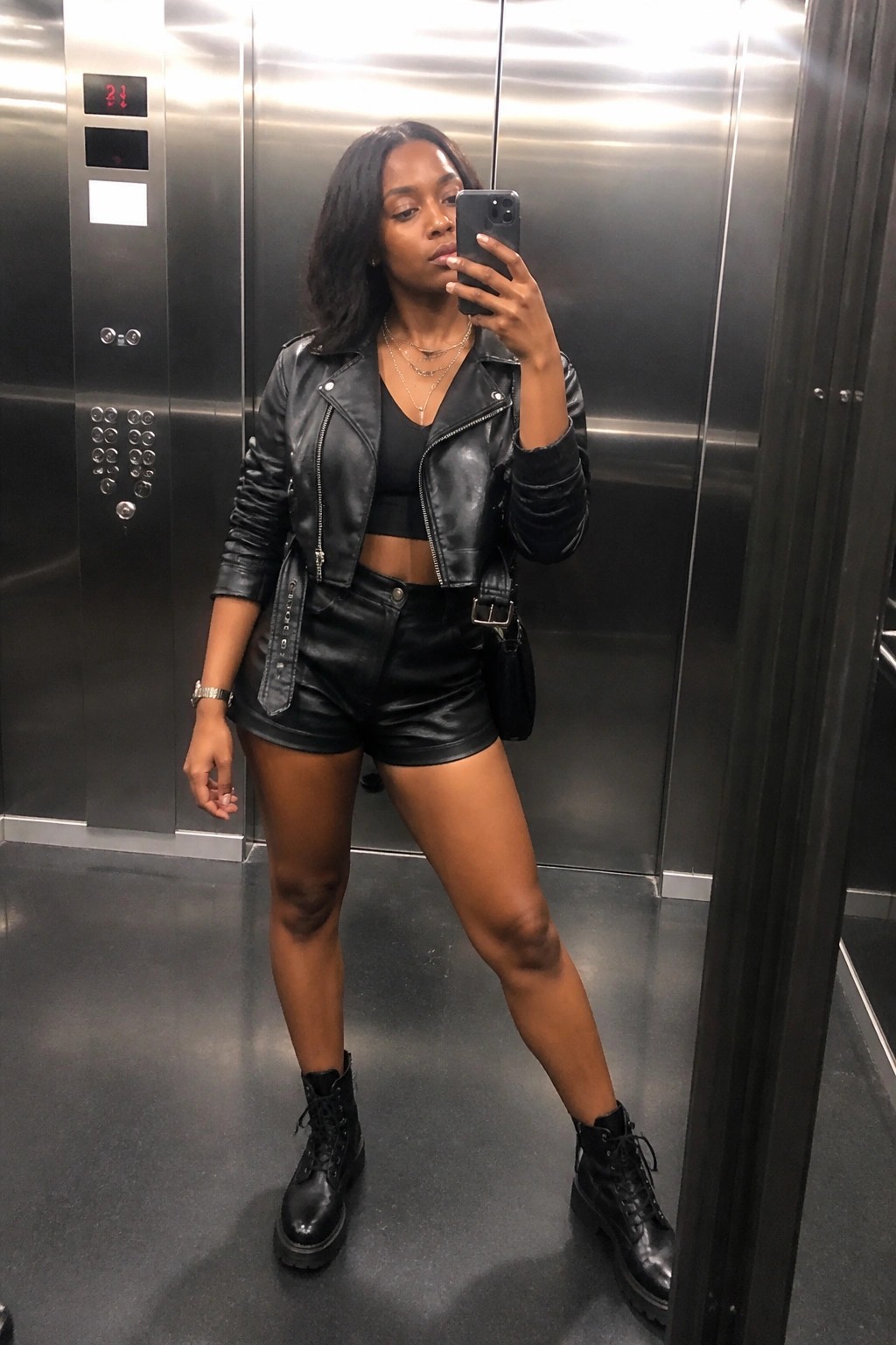 Woman wearing black leather moto jacket over black crop top with matching black leather shorts, black combat boots, layered necklaces, and small black bag in an elevator mirror selfie.