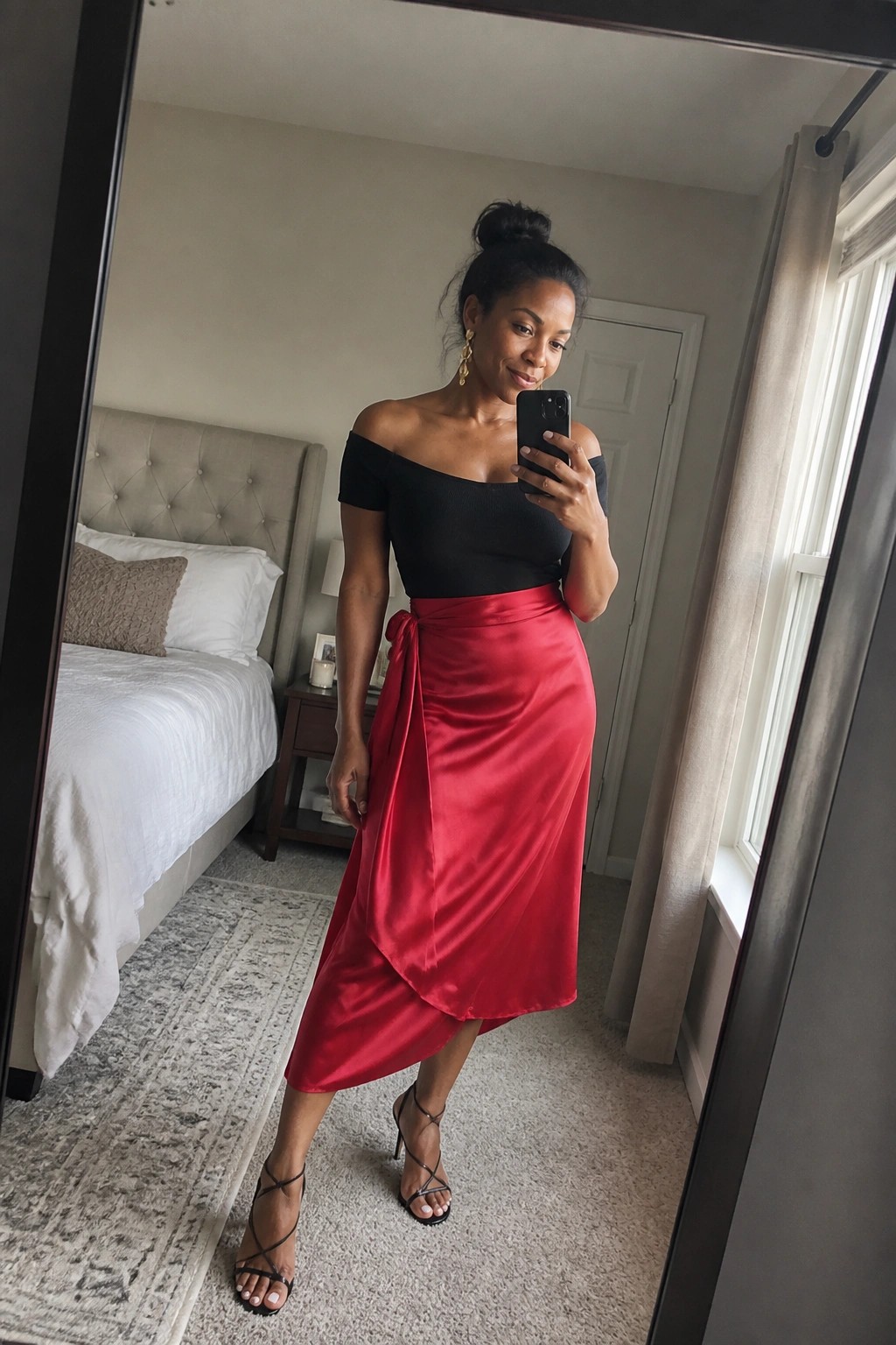 Woman in black off-shoulder top and red satin wrap skirt taking bedroom mirror selfie
