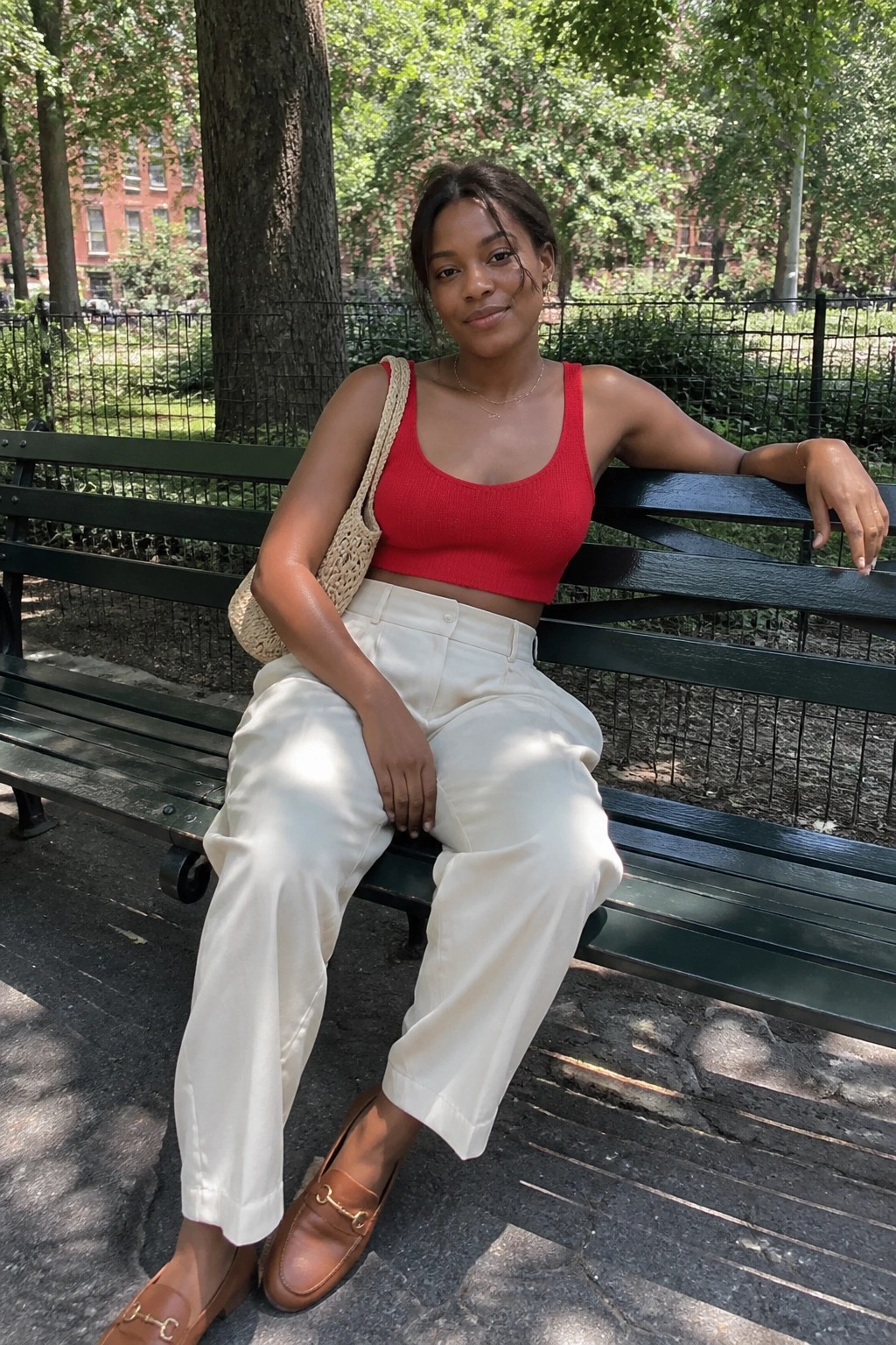 Woman in red crop top and white pants on green park bench with woven bag