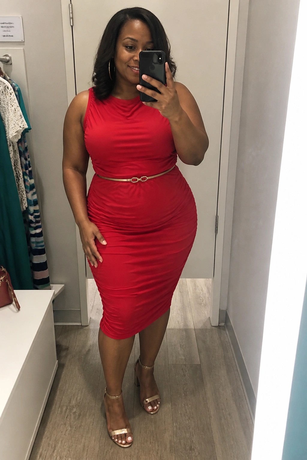 Woman in red ruched dress with gold belt taking mirror selfie in fitting room