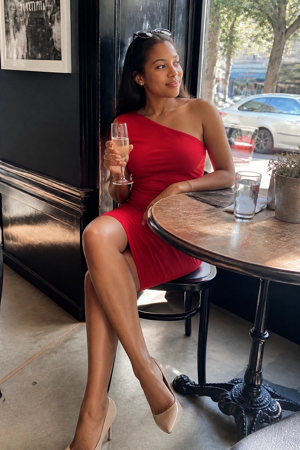 Woman in red one-shoulder dress holding champagne at cafe table with heels