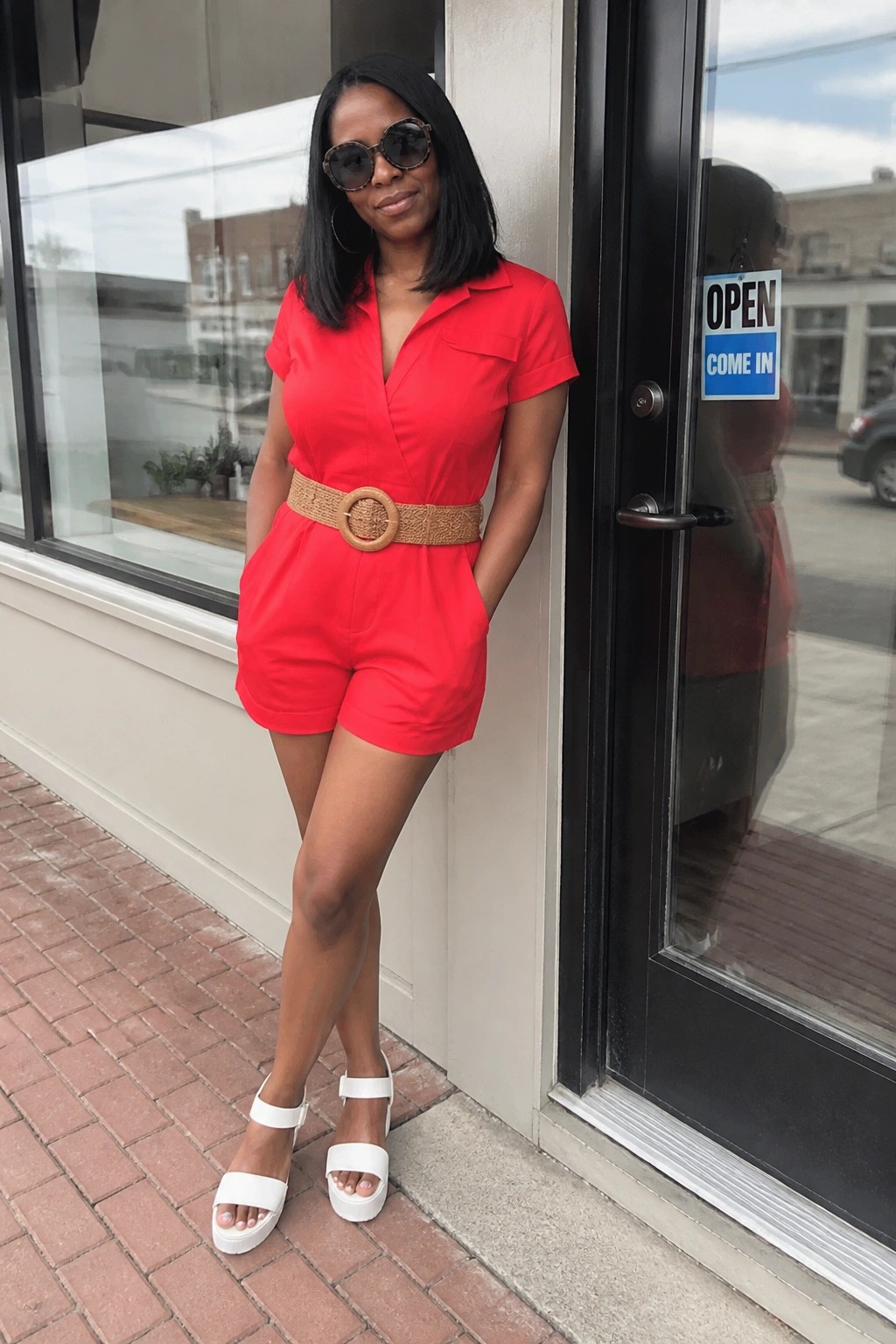 Woman in red romper with woven belt and white sandals at storefront door