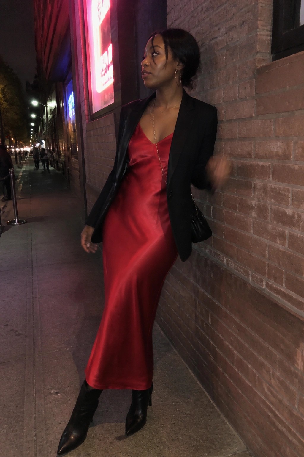 Woman wearing a long red satin slip dress layered under an oversized black blazer, paired with black pointed toe boots and gold jewelry including a long chain necklace.